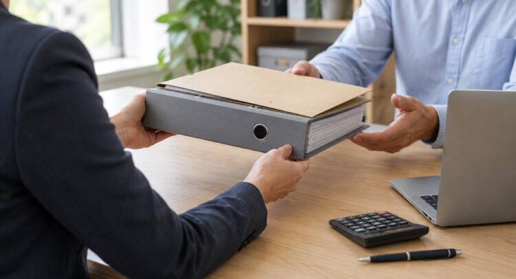 Deux personnes se passent un classeur gris et une chemise cartonnée au-dessus d'un bureau en bois, près d'un ordinateur portable et d'une calculatrice