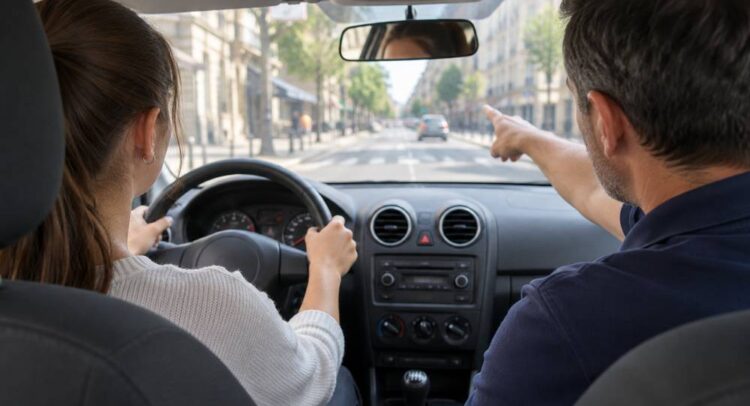 Depuis la banquette arrière d'une voiture-école, une jeune femme au volant regarde la rue que le moniteur, en polo bleu marine, lui montre du doigt